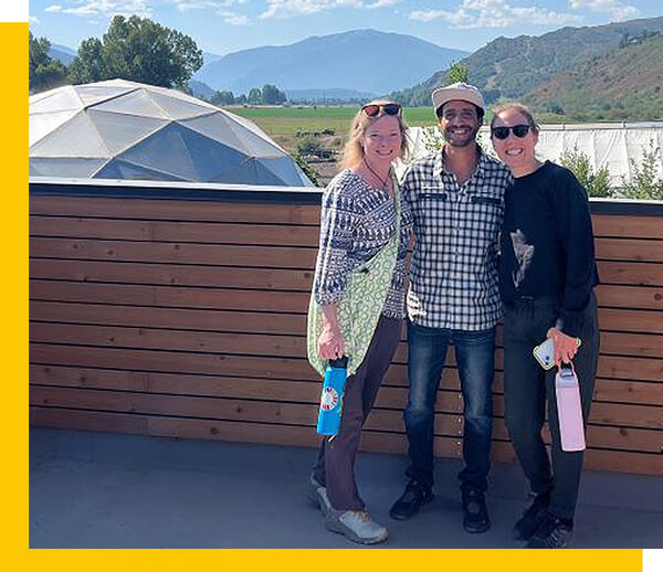 three group members pose for photo, mountains and a spherical green house in the background
